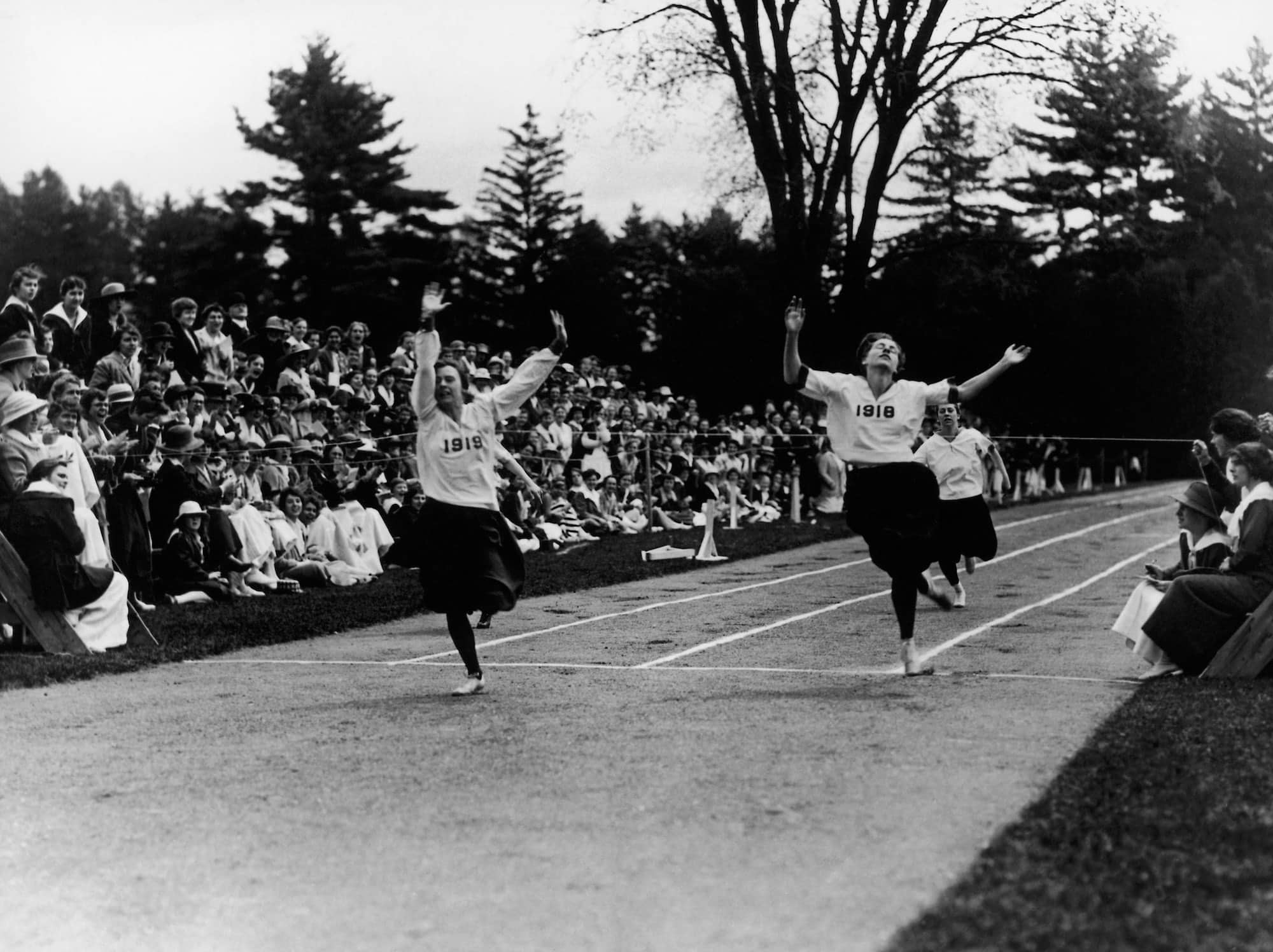 Historical black and white photo of two people wearing white sweatshirts with 1918 and 1919 imprinted on them, sprinting across the finish line of a dirt track with their hands in the air, while crowds of people seated at the sides of the track watch and cheer.